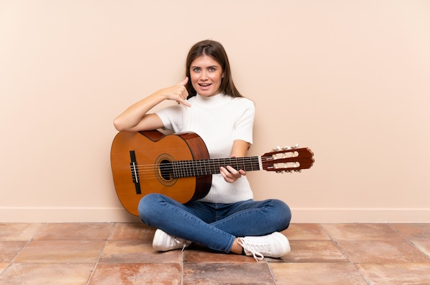 Młoda kobieta z gitary obsiadaniem na podłoga robi telefonu gestowi