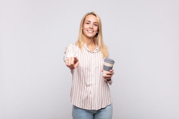 Młoda kobieta z kawą wskazującą na aparat z zadowolonym, pewnym siebie, przyjaznym uśmiechem, wybierająca Ciebie