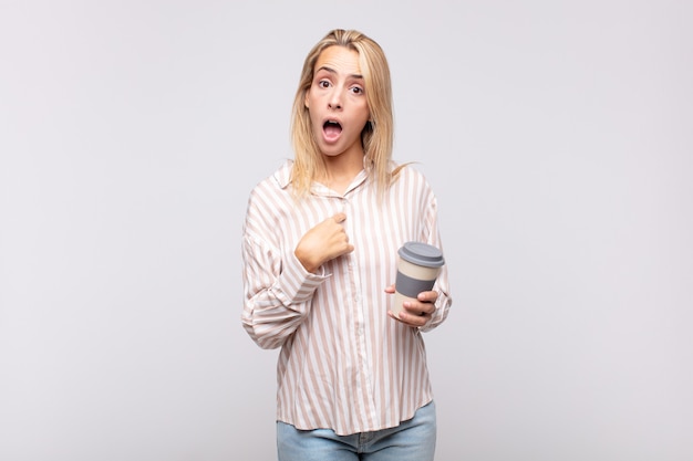 Młoda kobieta z kawą wyglądająca na zszokowaną i zaskoczoną z szeroko otwartymi ustami, wskazująca na siebie