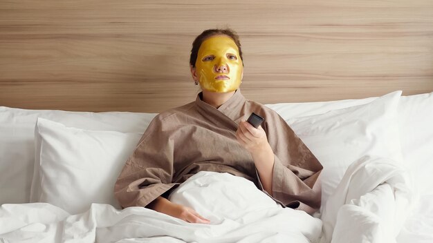 Młoda kobieta z kosmetyczną złotą maską na twarzy i pilotem do telewizora w ręku leży w miękkim łóżku w nowoczesnym pokoju hotelowym