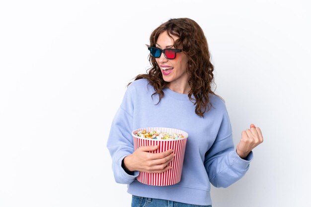 Młoda kobieta z kręconymi włosami na białym tle w okularach 3d i trzymająca duże wiadro popcornów, patrząc w bok