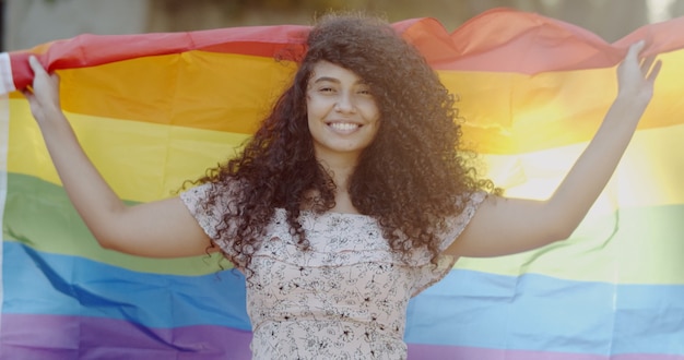młoda kobieta z kręconymi włosami przykrywająca flagę dumy lgbt trzymająca pięść w górze zakrywającą flagę lgbt;