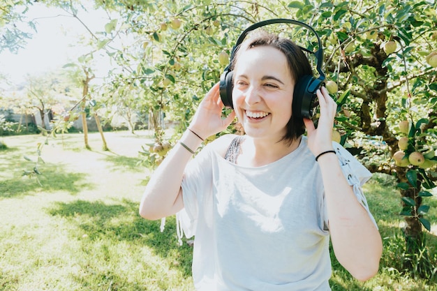 Młoda kobieta z krótkimi włosami bawi się słuchając muzyki za pomocą dużego zestawu słuchawkowego. Zajęcia zewnętrzne. Cieszyć się szczęśliwą młodą dziewczyną muzyki.