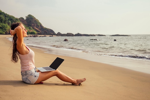 Młoda kobieta z laptopem na tropikalnych wakacjach, siedząc na piasku na bezludnej rajskiej wyspie na plaży nad morzem. Dziewczyna freelancer surfer na Oceanie Indyjskim.remote wolność pracy freelancing copyspace