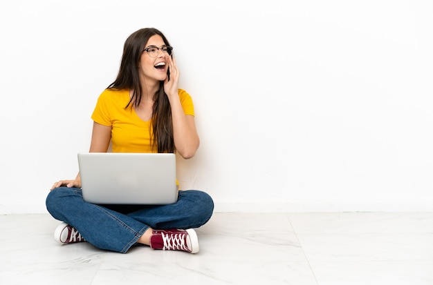 Młoda kobieta z laptopem siedząca na podłodze krzycząca z szeroko otwartymi ustami na boki