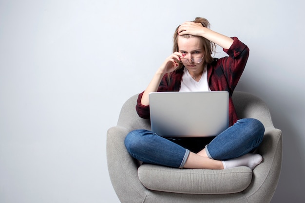Młoda kobieta z laptopem siedzi na miękkim wygodnym krześle i uśmiecha się, kobieta za pomocą komputera na białym pustym wal