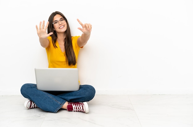 Młoda kobieta z laptopem siedzi na podłodze, licząc siedem palcami