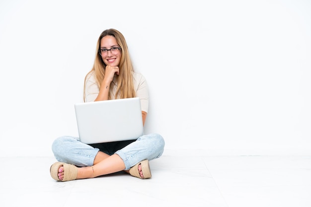 Młoda kobieta z laptopem siedzi na podłodze na białym tle w okularach i uśmiecha się