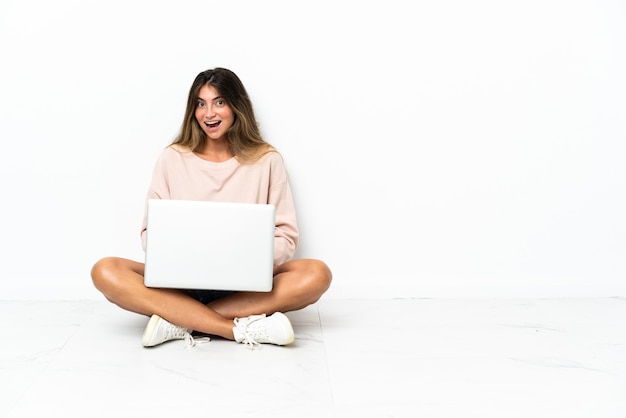 Młoda kobieta z laptopem siedzi na podłodze na białym tle z zaskoczeniem wyraz twarzy