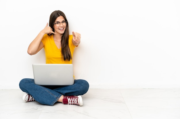 Młoda kobieta z laptopem siedzi na podłodze robi telefonowi gest i wskazuje przód