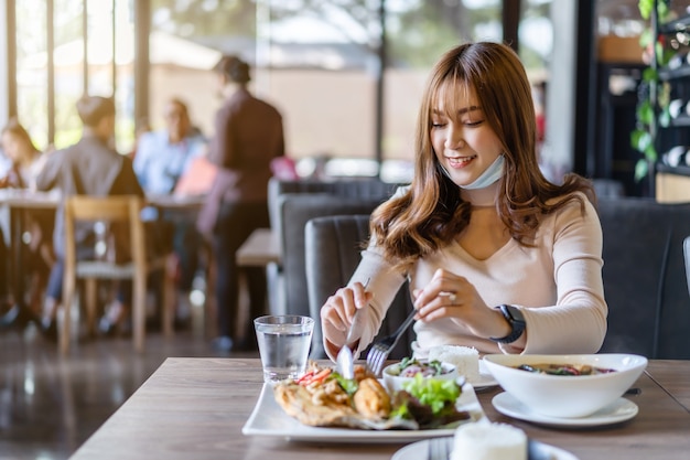 Młoda kobieta z maską na twarzy je jedzenie w restauracji, nowa normalna koncepcja ochrony przed pandemią koronawirusa (Covid-19)