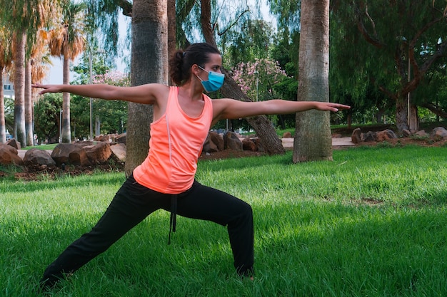 młoda kobieta z maską robi postawę jogi w parku w nowej normalności
