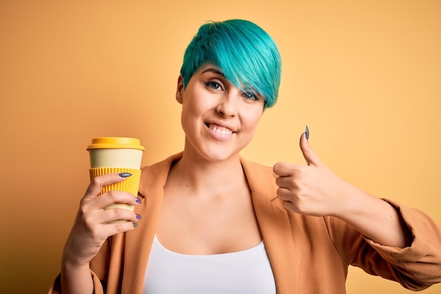 Młoda kobieta z niebieskimi modnymi włosami pije filiżankę kawy na żółtym odizolowanym tle zadowolona z dużym uśmiechem robi znak ok kciuk w górę palcami doskonały znak