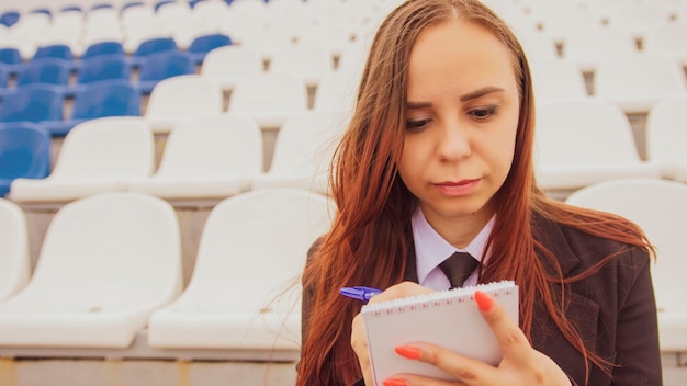 Młoda kobieta z notatnikiem, siedząca samotnie na trybunach stadionowych Dziennikarka zapisująca notatki podczas treningu sportowego na stadionie ulicznym