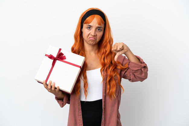 Młoda kobieta z pomarańczowymi włosami trzyma prezent na białym tle pokazując kciuk w dół z negatywnym wyrazem twarzy