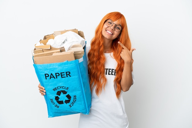 Młoda kobieta z pomarańczowymi włosami trzyma torbę do recyklingu pełną papieru do recyklingu na białym tle, wykonując gest kciuka w górę