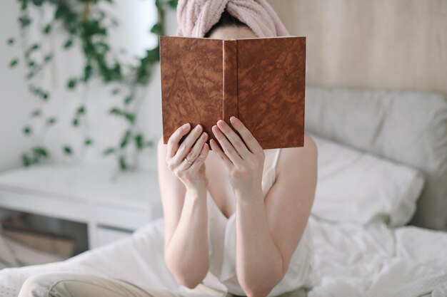 Młoda kobieta z ręcznikiem na głowie czytająca książkę rano w łóżku