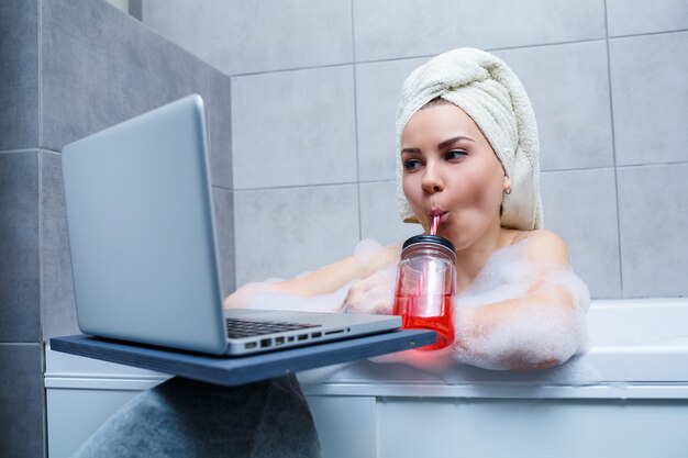 Młoda kobieta z ręcznikiem na głowie pije sok, oglądając film na laptopie, siedząc w wannie w salonie piękności. Zrelaksuj się w łazience bez pościeli. Koncepcja pielęgnacji ciała i relaksu.