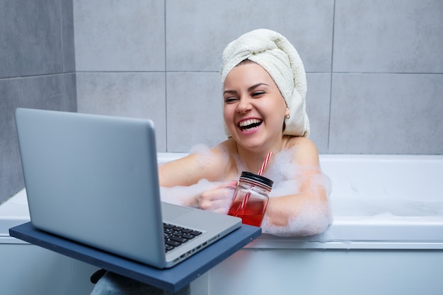 Młoda kobieta z ręcznikiem na głowie pije sok, oglądając film na laptopie, siedząc w wannie w salonie piękności. Zrelaksuj się w łazience bez pościeli. Koncepcja pielęgnacji ciała i relaksu.