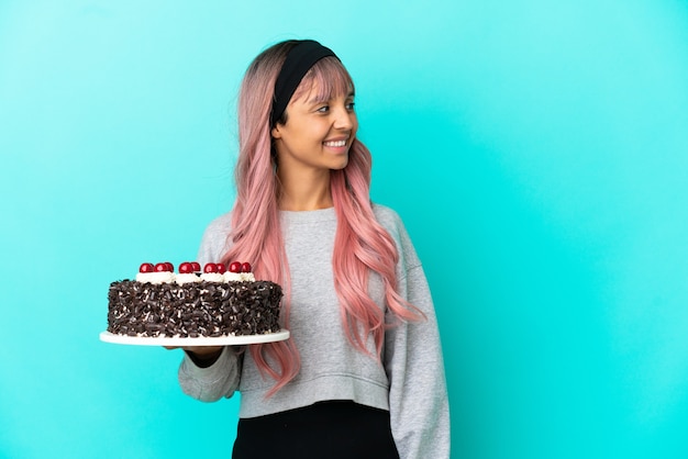 Młoda kobieta z różowymi włosami trzyma tort urodzinowy na białym tle na niebieskim tle, patrząc w bok i uśmiechając się