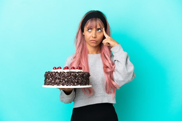 Młoda kobieta z różowymi włosami trzymająca tort urodzinowy na białym tle na niebieskim tle myśląca o pomyśle