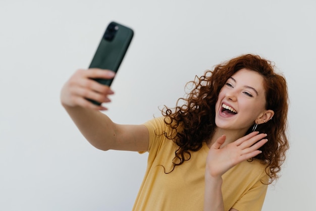 Młoda kobieta z rudymi włosami robi selfie lub wideorozmowę na białym tle