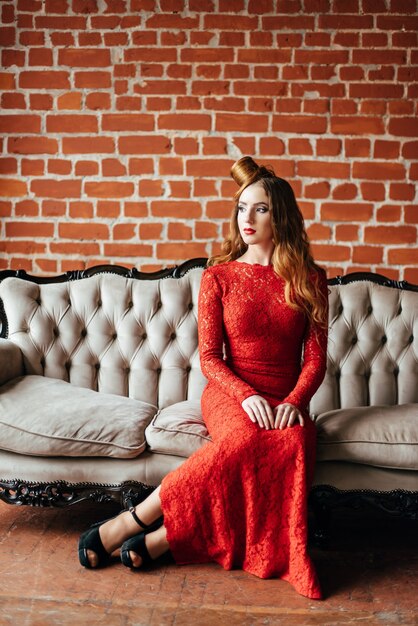 Młoda kobieta z rudymi włosami w jaskrawoczerwonej sukience w jasnym pokoju na zabytkowej sofie