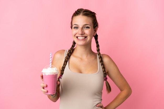 Młoda kobieta z truskawkowym koktajlem na białym tle na różowym tle pozowanie z rękami na biodrze i uśmiechnięty