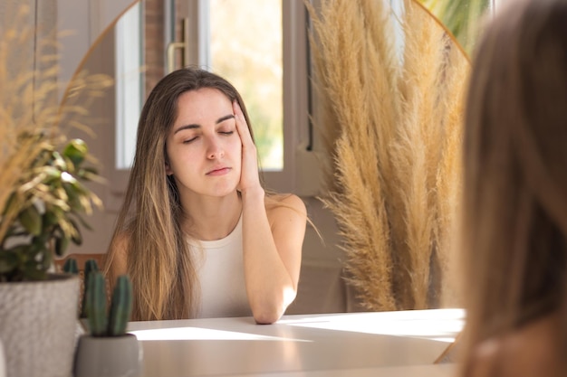Młoda kobieta z zamkniętymi oczami siedząca przed lustrem z bólem głowy