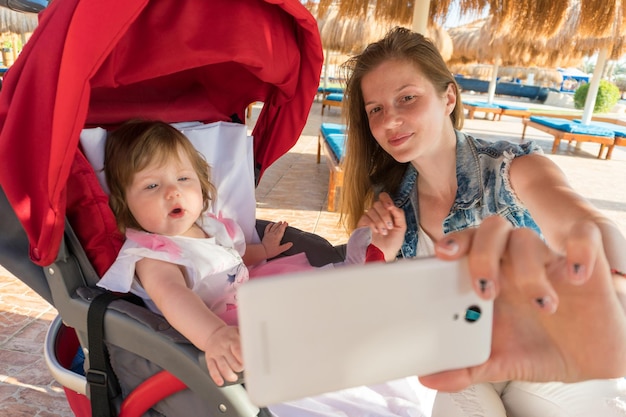 Młoda kobieta za pomocą smartfona, aby zrobić selfie w pobliżu wózka z uroczą córeczką w słoneczny dzień w ośrodku na wakacjach