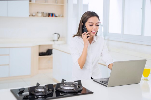 Młoda kobieta za pomocą telefonu komórkowego i laptopa w kuchni