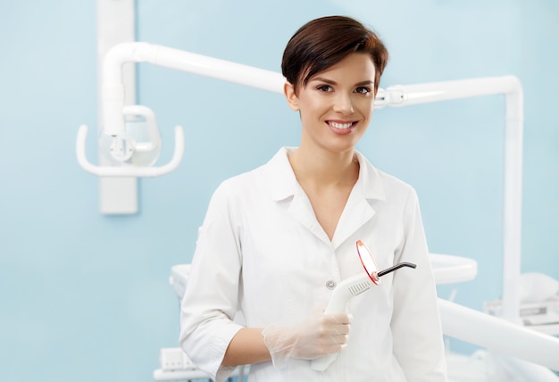 Młoda kobiety lekarka w dentysty biurze. Piękna uśmiechnięta kobieta trzyma błękitnego stomatologicznego leczy światło w białym lab żakiecie. Klinika dentystyczna. Stomatologia