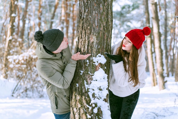 Młoda kochająca para wygląda zza drzewa i przesyła sobie buziaka w zaśnieżonym lesie Mężczyzna i kobieta w słonecznym zimowym parku