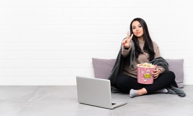 Młoda Kolumbijska dziewczyna trzyma puchar popcorns i pokazuje film w laptopie uśmiecha się znak zwycięstwa i pokazuje