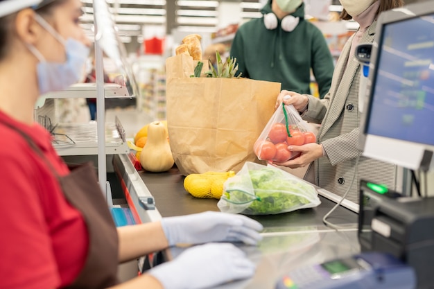 Młoda konsumentka stawiająca świeże pomidory i inne produkty na ladzie, stojąc przy kasie przed kasjerem w supermarkecie