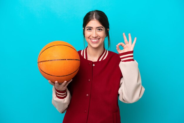 Młoda koszykarz kaukaski kobieta na białym tle na niebieskim tle pokazując znak ok palcami