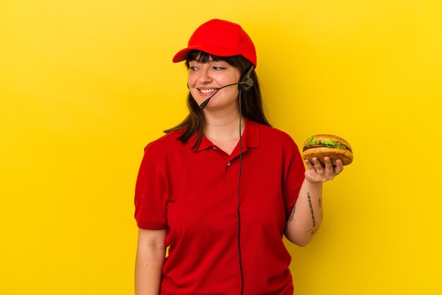 Zdjęcie młoda krągła kobieta kaukaska trzymająca burgera na żółtym tle wygląda na bok uśmiechniętą, wesołą i przyjemną.