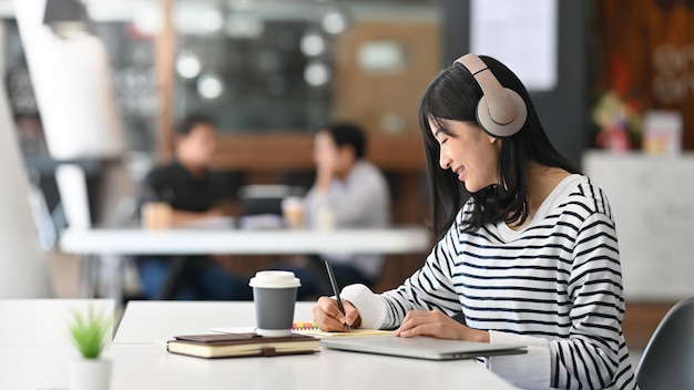 Młoda kreatywnie kobiety writing i słuchająca muzyka w nowożytnym biurze