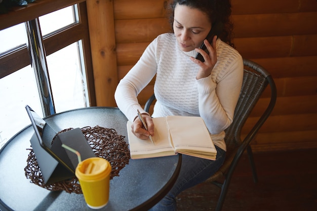 Młoda, kręcona kobieta skupiona w pracy, pisząc w dzienniku, rozmawiając przez smartphoneon, siedząc przy oknie w kreatywnej drewnianej kawiarni