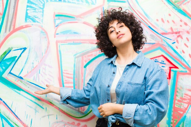 Młoda ładna afro kobieta czuje się szczęśliwa i wesoła, uśmiecha się i wita, zapraszając przyjaznym gestem do ściany graffiti