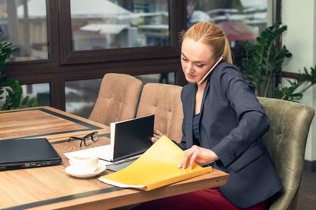 Zdjęcie młoda ładna biznesowa kobieta opowiada na telefonie i pracuje z dokumentami przy biurem.
