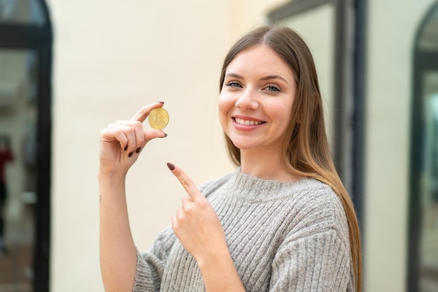 Młoda ładna blondynka trzymająca bitcoina na zewnątrz i wskazująca go