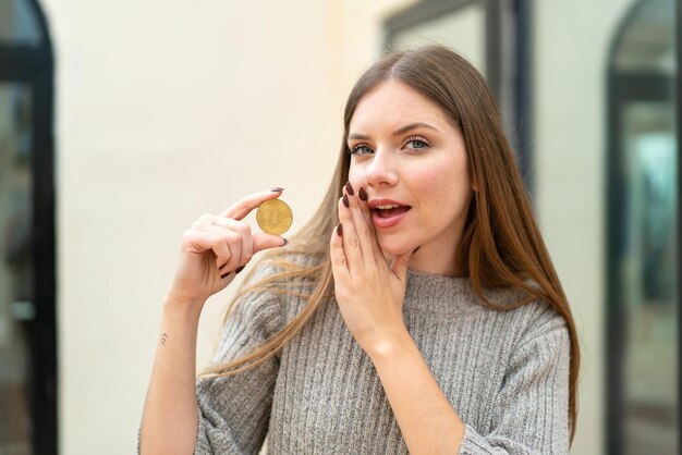 Młoda ładna blondynka trzymająca Bitcoina na zewnątrz, szepcząca coś