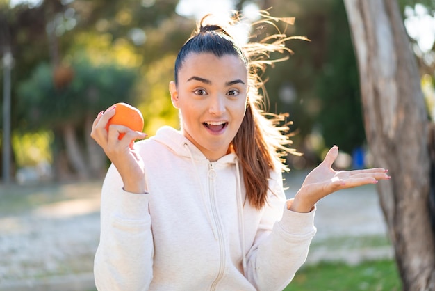 Młoda ładna brunetka trzyma pomarańczę na zewnątrz z zszokowanym wyrazem twarzy