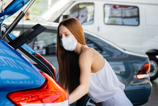 Młoda ładna dziewczyna podczas pandemii ogląda nowy samochód w salonie samochodowym w masce. Sprzedaż i kupno samochodów w okresie pandemii.