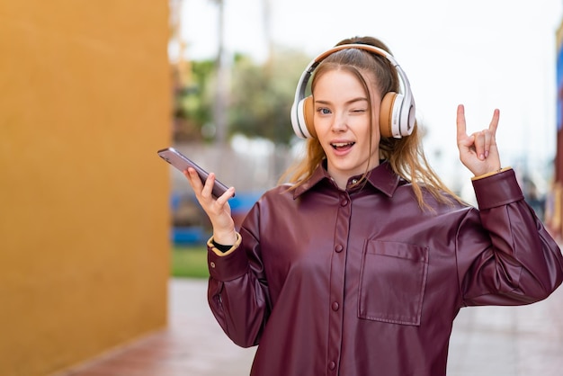 Młoda ładna dziewczyna słucha muzyki na zewnątrz za pomocą telefonu komórkowego, wykonując rockowy gest