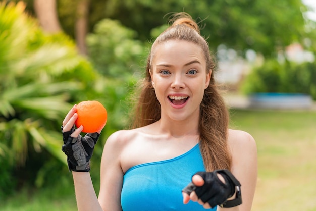 Młoda ładna dziewczyna sportowa trzyma pomarańczę na zewnątrz zaskoczona i wskazująca przód