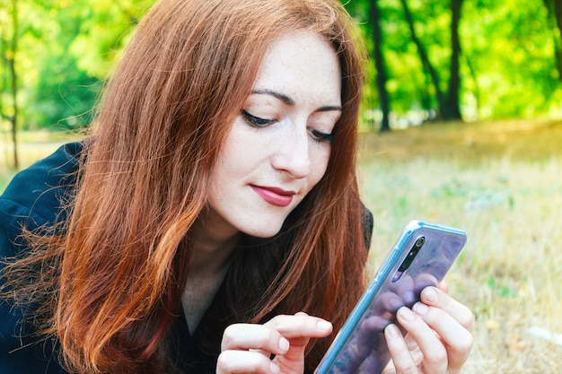 Młoda ładna dziewczyna z rudymi włosami, patrząc na telefon w lecie.