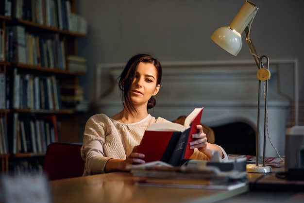 Młoda ładna i entuzjastyczna studencka dziewczyna czyta książkę w bibliotece uniwersyteckiej pod lampą stołową