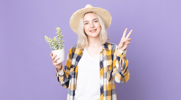 Młoda ładna kobieta albinos uśmiechnięta i wyglądająca przyjaźnie, pokazująca numer dwa i trzymająca kaktusa z rośliną doniczkową